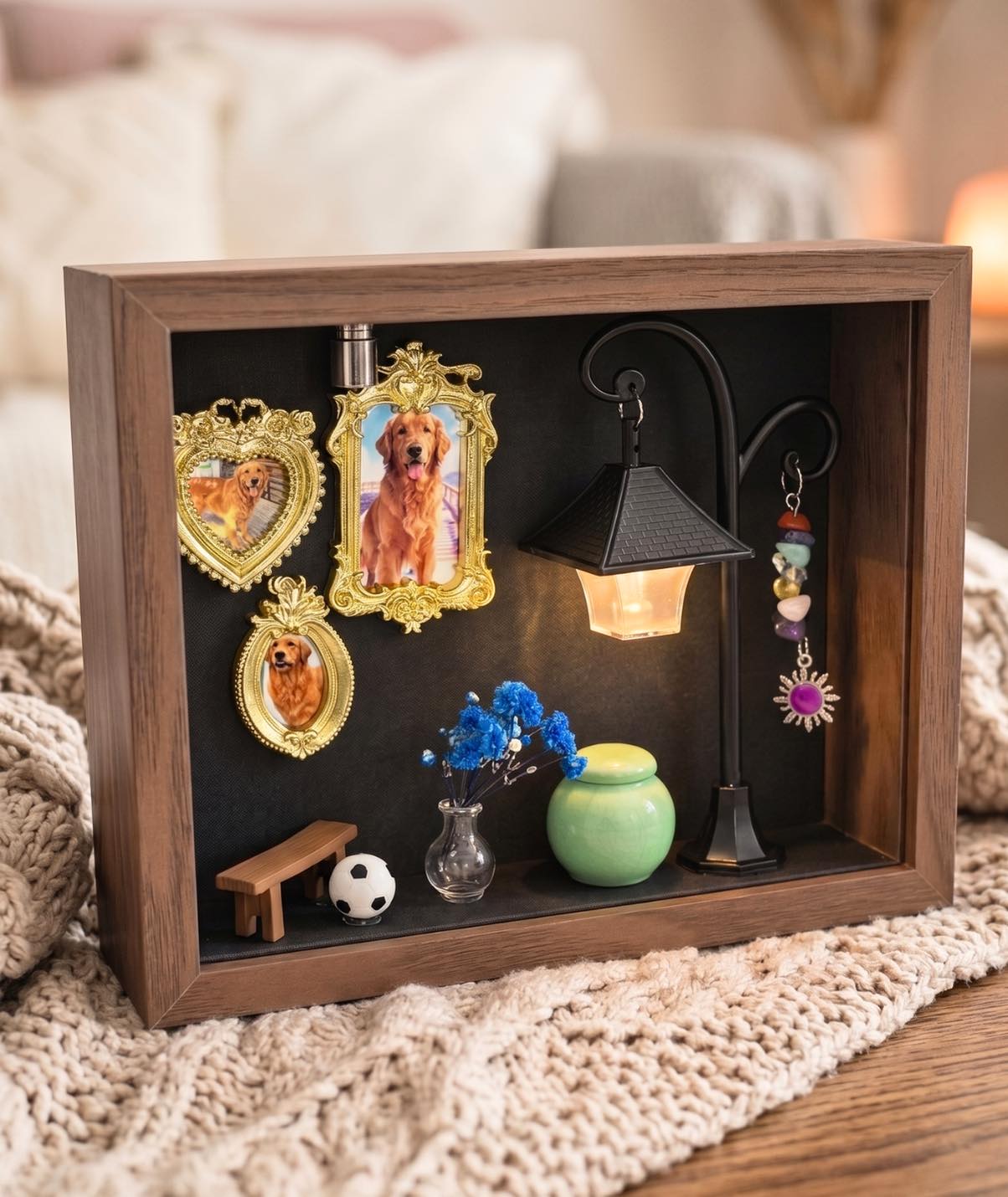 A customized wooden 3D pet memorial shadow box displayed on a cozy knitted blanket in a warm home setting. The diorama inside features vintage framed photos of a Golden Retriever, an illuminated miniature street lamp with a hanging Natural Amethyst charm, blue miniature flowers, a tiny park bench, a small soccer ball, and a green ceramic keepsake jar.