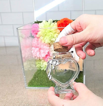 Hands holding a personalized heart-shaped glass pet fur keepsake bottle with a cork stopper.