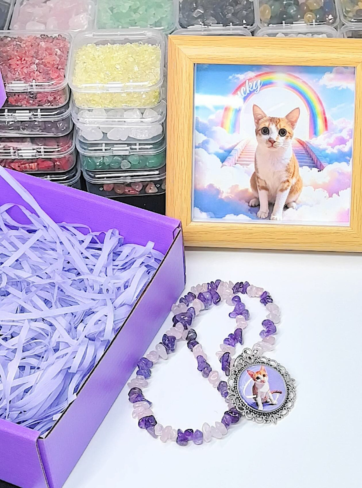 A custom pet photo necklace with natural amethyst and rose quartz beads displayed next to a purple gift box and a framed remembrance photo of a cat on a rainbow bridge.