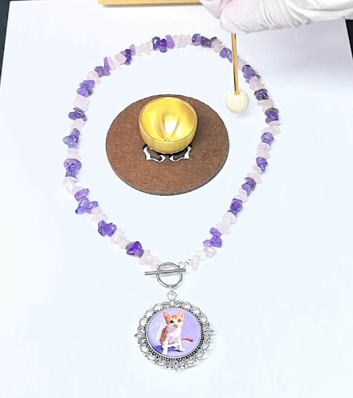 A flat lay of a personalized pet photo keepsake necklace made with natural amethyst and rose quartz beads, arranged around a small brass singing bowl.