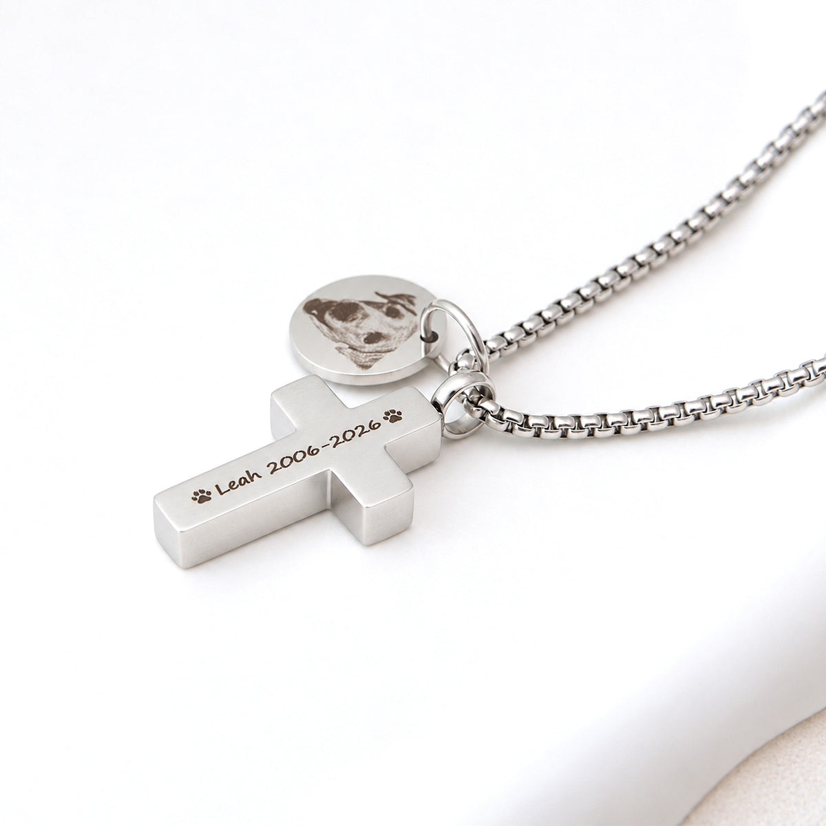 Close-up of a silver stainless steel pet memorial cross necklace engraved with a dog's name and dates, beside a round photo charm.
